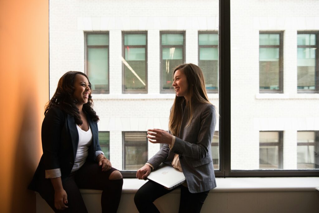 Two women sitting facing each other and talking in front of a large window.